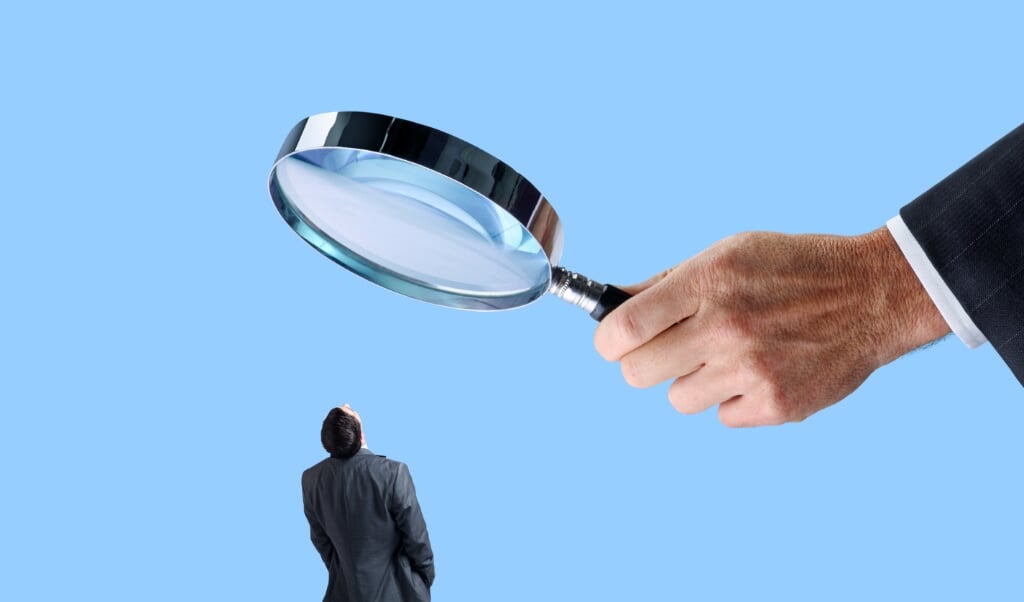A businessman looks up at a large magnifying glass that is being held by an equally large hand isolated on a blue background.  (beeld iStock)