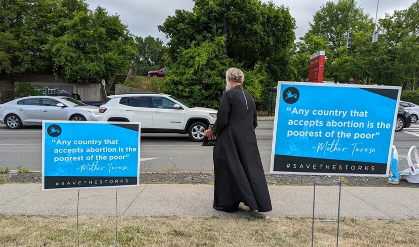 Langs de drukke, doorgaande weg in Troy demonstreert de Russisch-orthodoxe priester Michael Fritz. Roerloos staat hij op de stoep, een afwezige glimlach op zijn gezicht, de blik strak naar beneden.