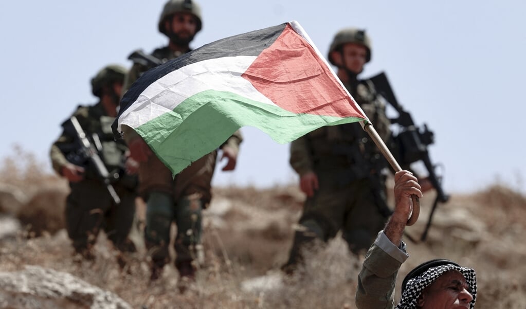Een Palestijnse man met de Palestijnse vlag terwijl Israëlische veiligheidstroepen toekijken. (beeld jaafar Ashtiyeh / afp)