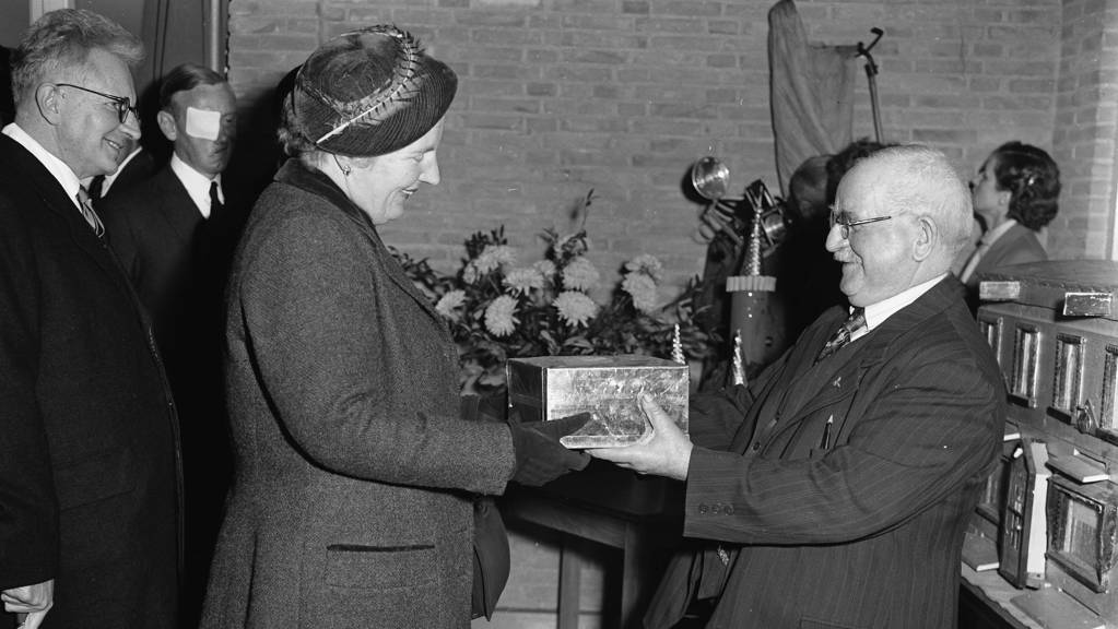 Tijdens haar werkbezoek aan Emmen in 1958 ontvangt koningin Juliana een blikken trommel met raathoning van de tachtigjarige imker J. Jeuring.