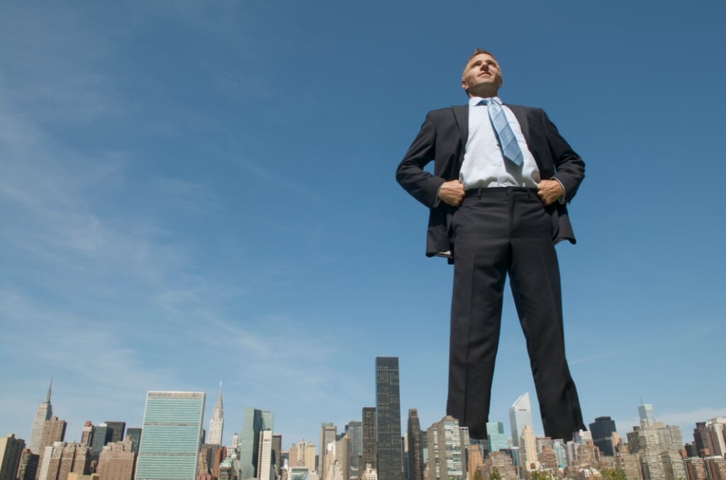 Confident smiling tall businessman giant standing with his hands on his hips, towering over the city skyline  (beeld Getty Images/iStockphoto)