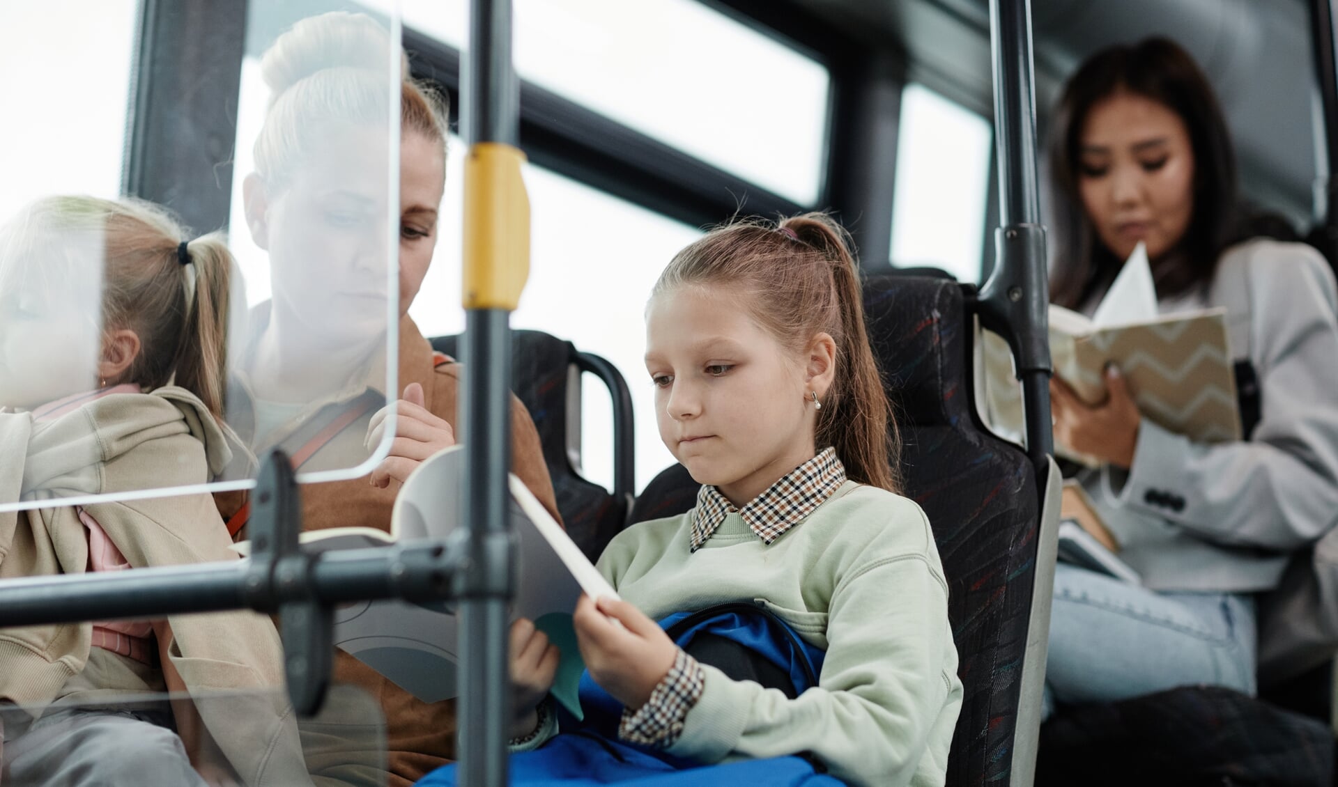 Portrait of schoolgirl reading book on bus while traveling by public transport with mom in city