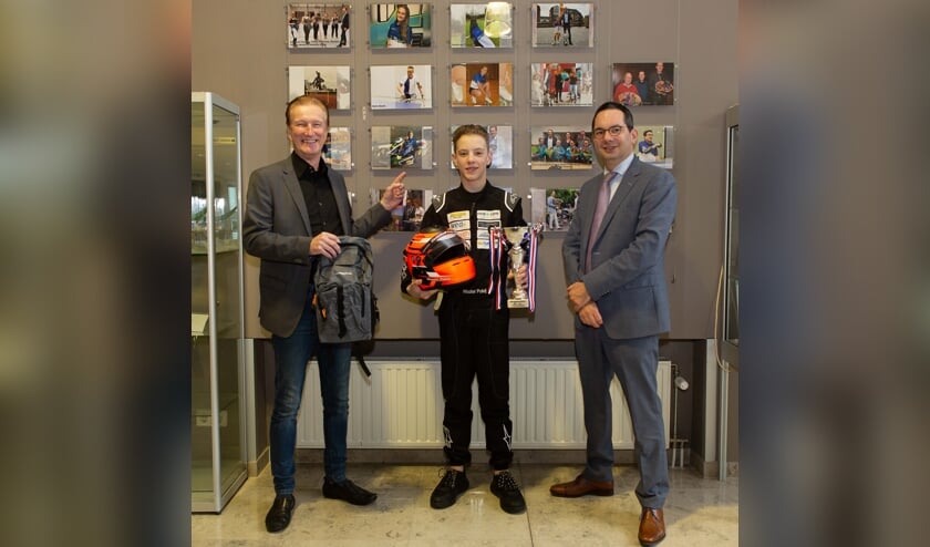 Met de gewonnen beker, zijn helm en rugtas voor de fotowand van alle winnaars. Wethouder Hans de Waal wijst de foto van Wouter aan.