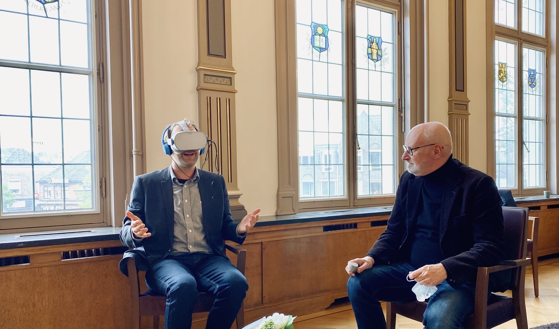 Wethouder Roozendaal probeert de VR-bril uit, vrijwilliger Marcel kijkt toe.