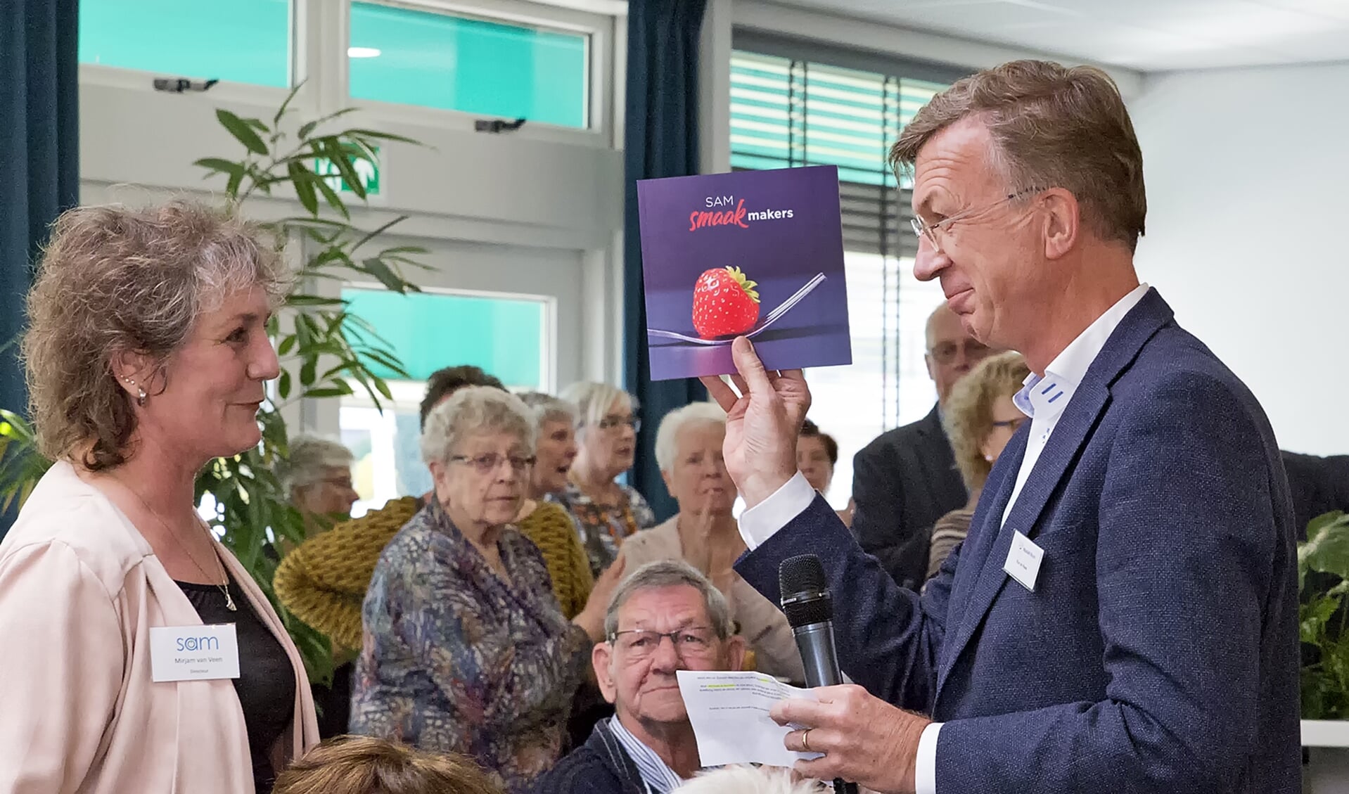 Ron de Haas, directeur Mozaïek Wonen, ontvangt uit handen van Mirjam van Veen, directeur SAM, het eerste exemplaar van het kookboekje dat de medewerkers van SAM hebben samengesteld