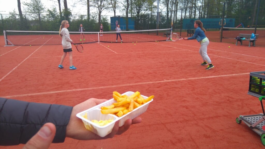 De tennisleraar is alvast aan het bakje patat begonnen, terwijl de kids nog druk bezig zijn met de oefeningen.