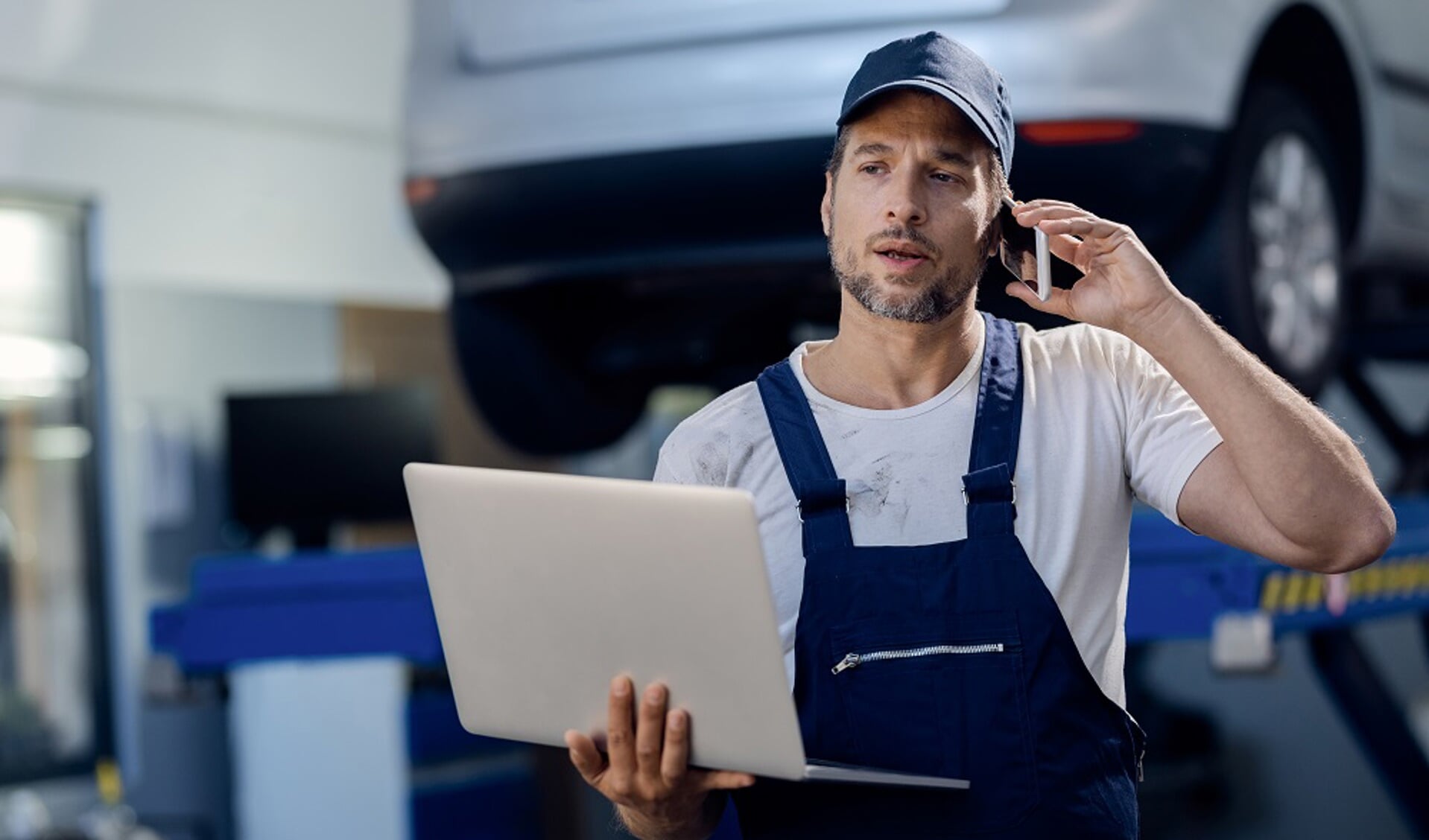 Mid adult mechanic using computer and talking on mobile phone at auto repair shop.