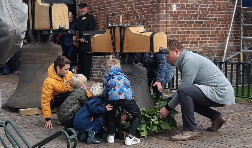 <p>Gedeputeerde Roy de Witte (rechts) legt samen met Tineke (achter de klok) en een paar kinderen een krans om de kleine klok, als 'welkom terug'. Op de achtergrond 'Klepper Derk' die iedereen naar de kerk haalde.</p>