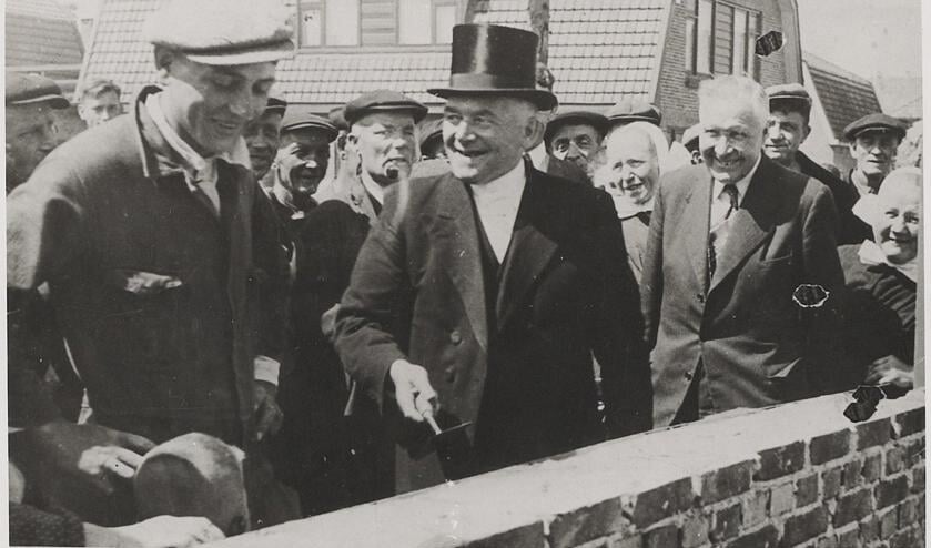 <p>De eerste steen wordt gelegd aan de Bevervoorde door ds. J. (Jan) van Wier. Aannemer Gerhard Pas (Gerrad van 'n Roes) kijkt toe, daarachter andere gemeenteleden.</p>