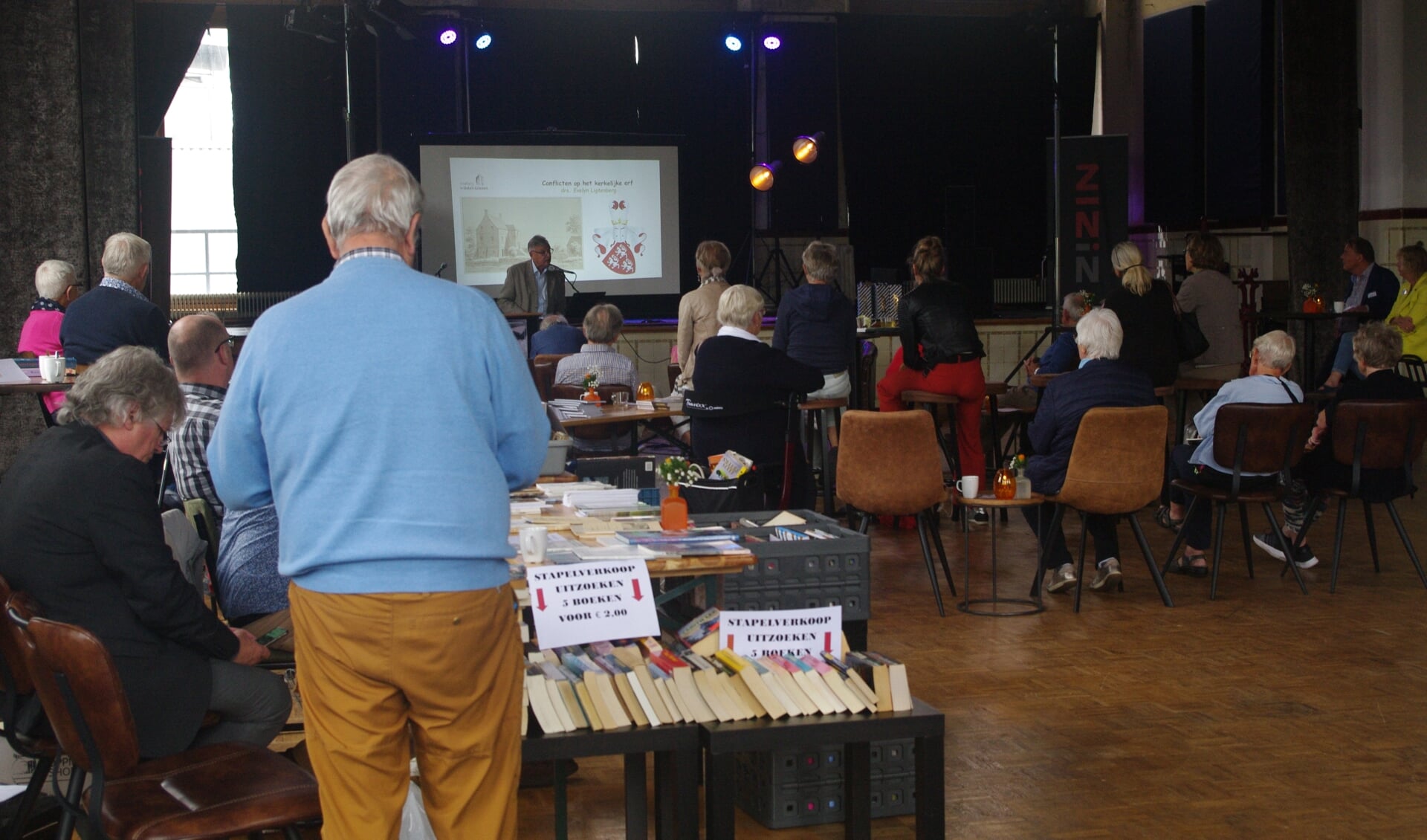 Diverse schrijvers en verzamelaars verkopen (en signeren) hun boeken tijdens de boekenmarkt van het Schrijversfestival, terwijl in een andere ruimte lezingen worden gegeven (hier in De Smidse, 2021).