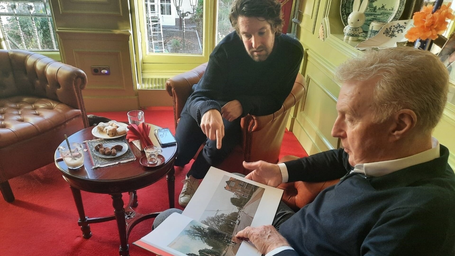 André van Duin bladert met onze verslaggever Peter van Drunen door het boek ‘Groeten uit Rotterdam’ met foto’s van nu en honderd jaar geleden. “Kijk, dit is Delfshaven. Hier heb ik veel gespeeld.” Foto: Rico Hop