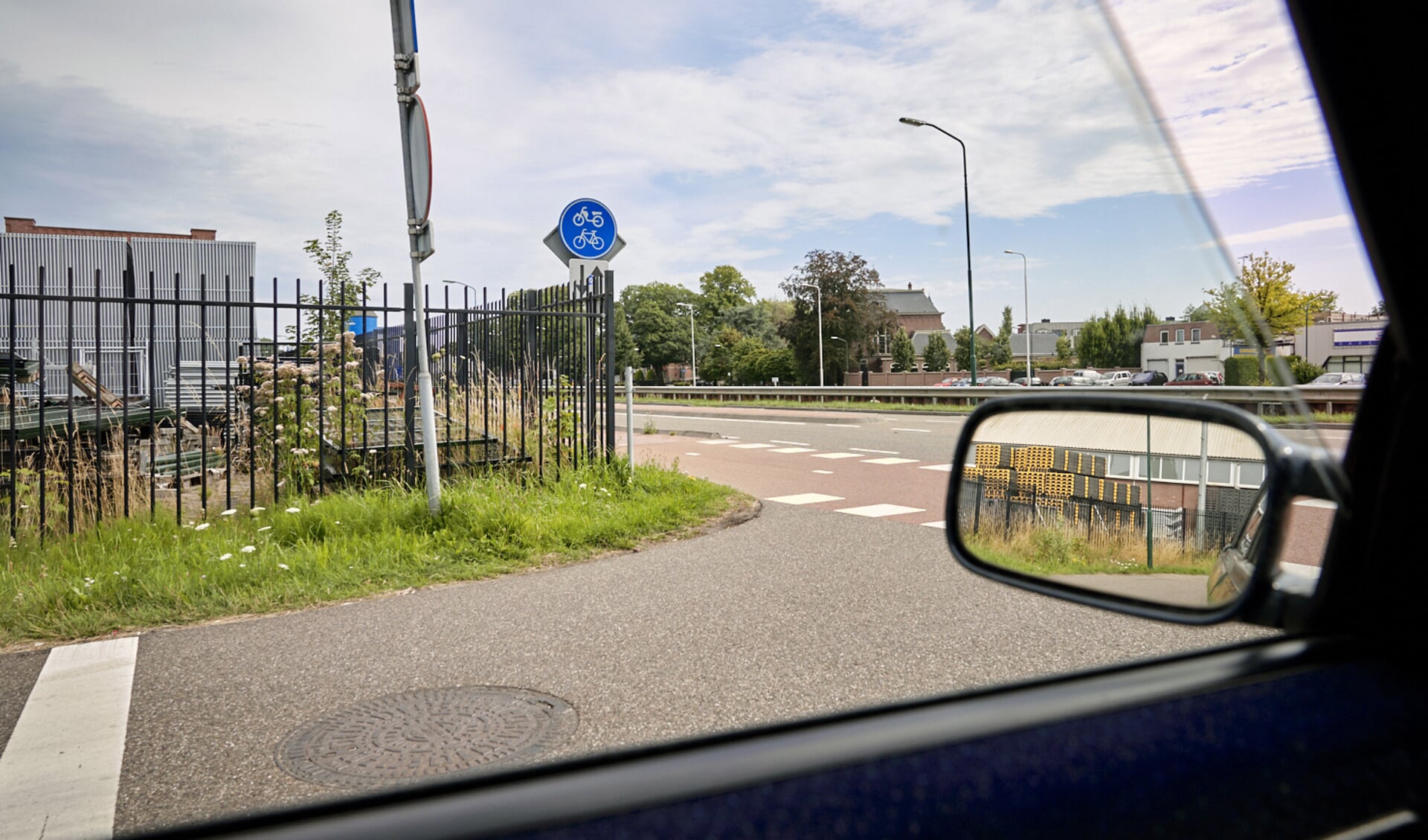 Het zicht van een afdraaiende auto vanuit de Wellestraat richting de Bosscheweg. Voordat de auto de weg op kan, moet het eerst nog voorbij een fietspad, dat beperkt zichtbaar is door de hekwerken aan beide zijden