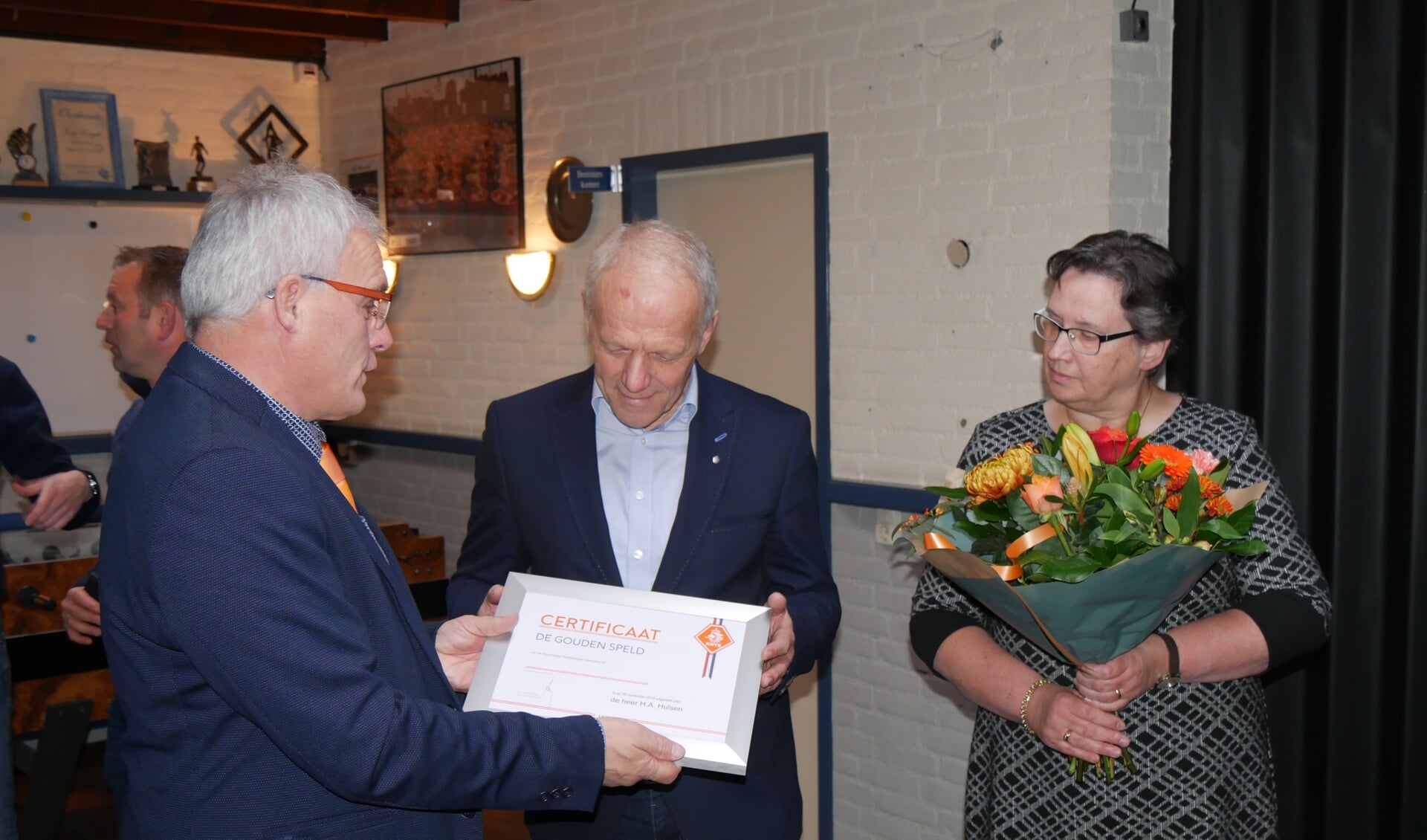 Henk ontvangt het certificaat behorende bij de gouden speld uit handen van Toon Kerkhof, met naast hem zijn vrouw Marieke
