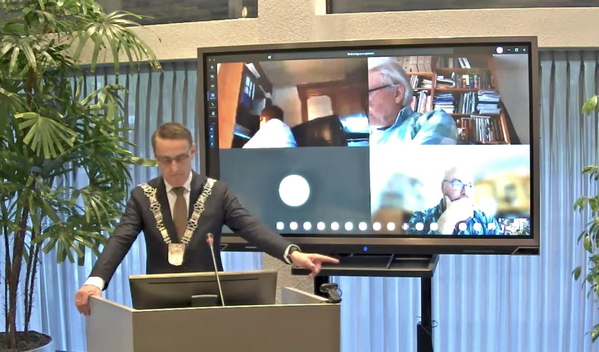 Burgemeester Van der Meijden praat de gemeenteraad bij. Op het scherm enkele raadsleden in beeld die vanuit eigen huis meeluisteren en vragen kunnen stellen