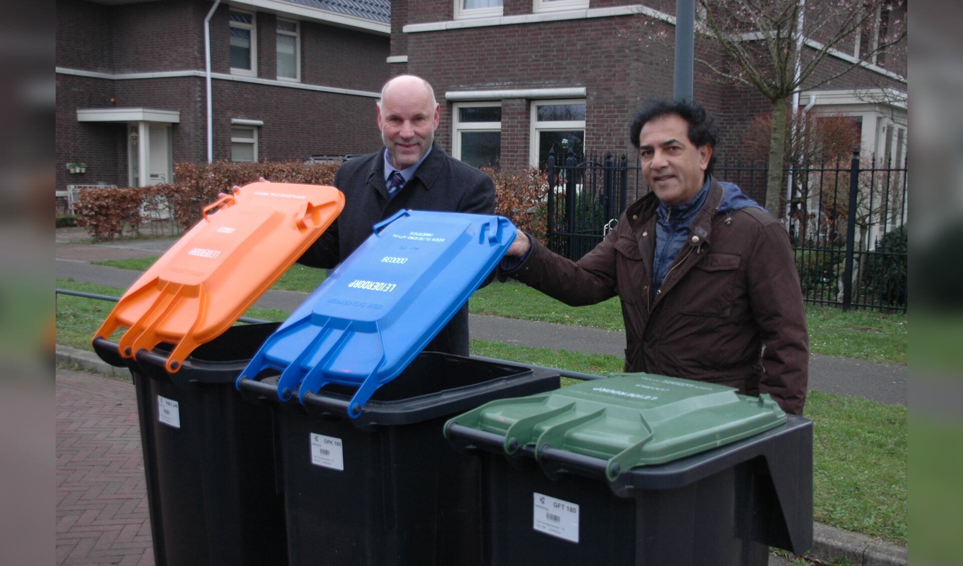 Wethouder McDaniel (links) heeft de eerste set minicontainers overhandigd aan bewoner Rob Mohanlal. De groene container is voor GTF, de blauwe voor papier en de oranje voor PMD.  | Foto: Corrie van der Laan