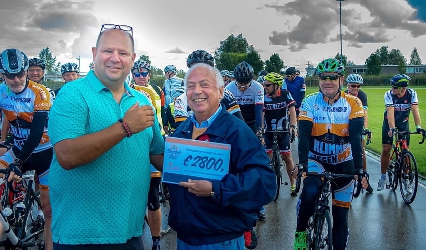 Paco Busteros León (links) overhandigt de cheque met de opbrengst over 2018 aan een vertegenwoordiger van het Instituut Asbestslachtoffers. Op de achtergrond staan de deelnemers aan de toertocht klaar voor de start.