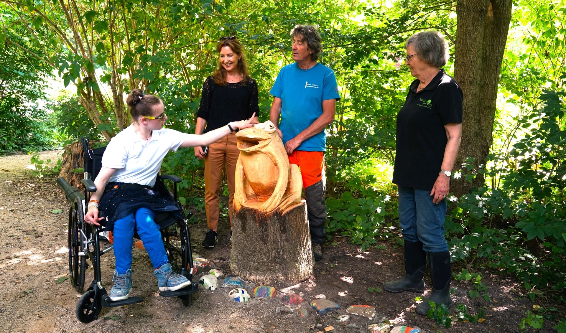 Nadja - naar wie Stichting Nadja is genoemd - aait de gloednieuwe houten kikker terwijl haar moeder Anne van Zwieten, boomkunstenaar James Arbor en voorzitter Riny Vons van de natuurspeeltuin toekijken.
