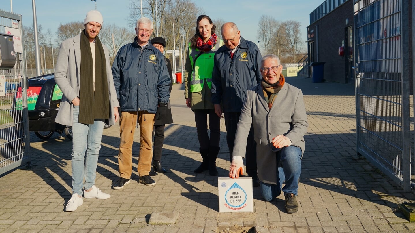 Wethouder Herman Romeijn toont de tegel die hij even later op zijn plek naast de put legt. Rotarians Bart Kerner en Pierre van Overloop, manager Ferdi Dreef van Sportfondsen Leiderdorp en Allison Bartlet van het MEC kijken toe.