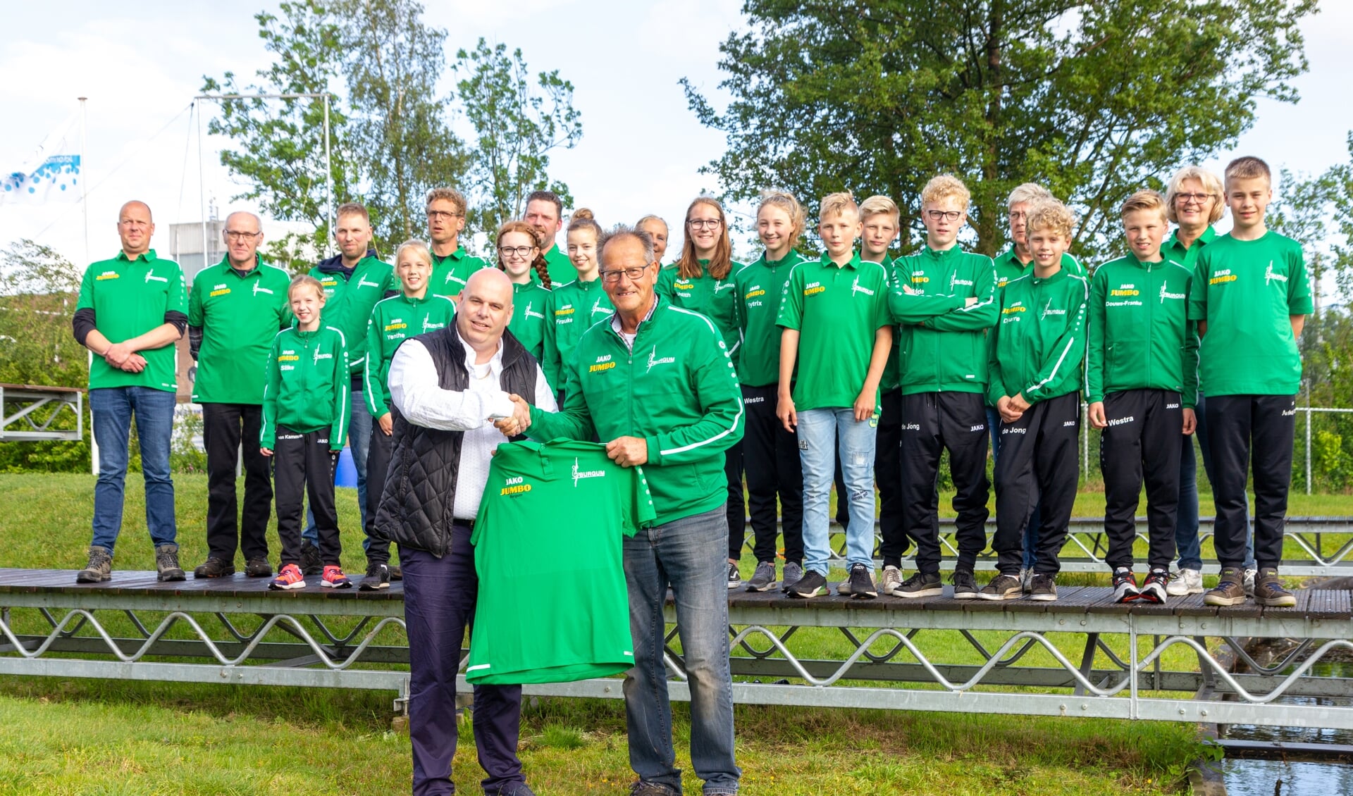Johan Postma, bedrijfsleider van Jumbo Burgum (links voor), overhandigt het nieuwe clubshirt aan Hans Helmholt, voorzitter van Ljeppersclub Burgum. Op de schans staan leden en vrijwilligers van de club.