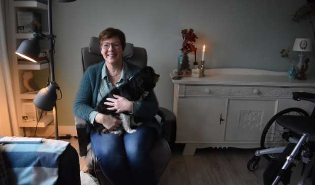 Pietie Dijkstra met haar hondje en rechts haar triple-rolstoel voor in huis. 