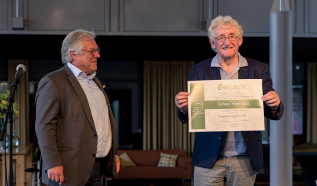 <p>Johan Veenstra toont de oorkonde die hij net uit handen kreeg van wethouder Robin Hartogh Heys van Smallingerland.</p>