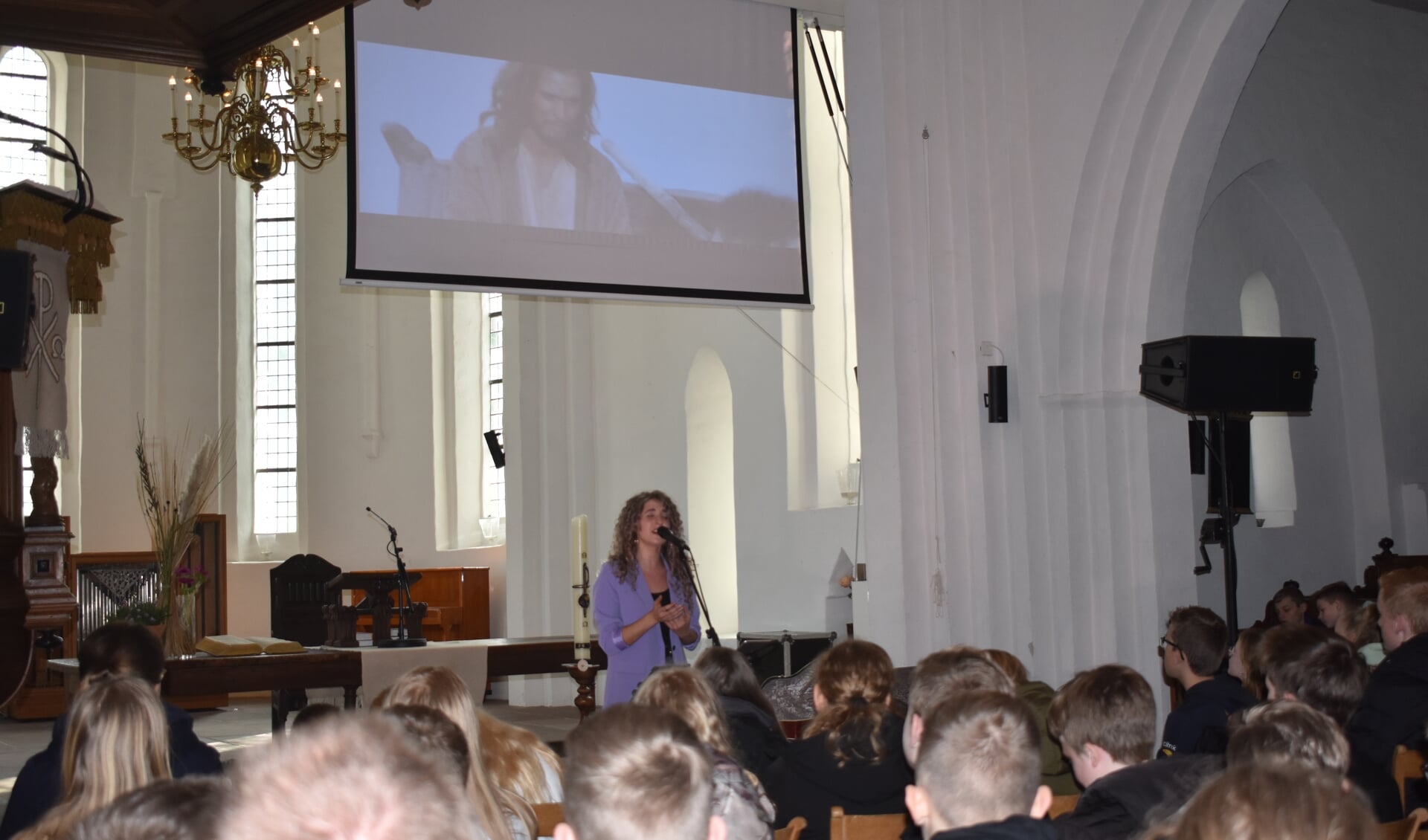 Sera Noa in de Kruiskerk in Burgum, onder een speelfilmbeeld  van Jezus, zingt voor leerlingen van CSG Liudger Burgum.