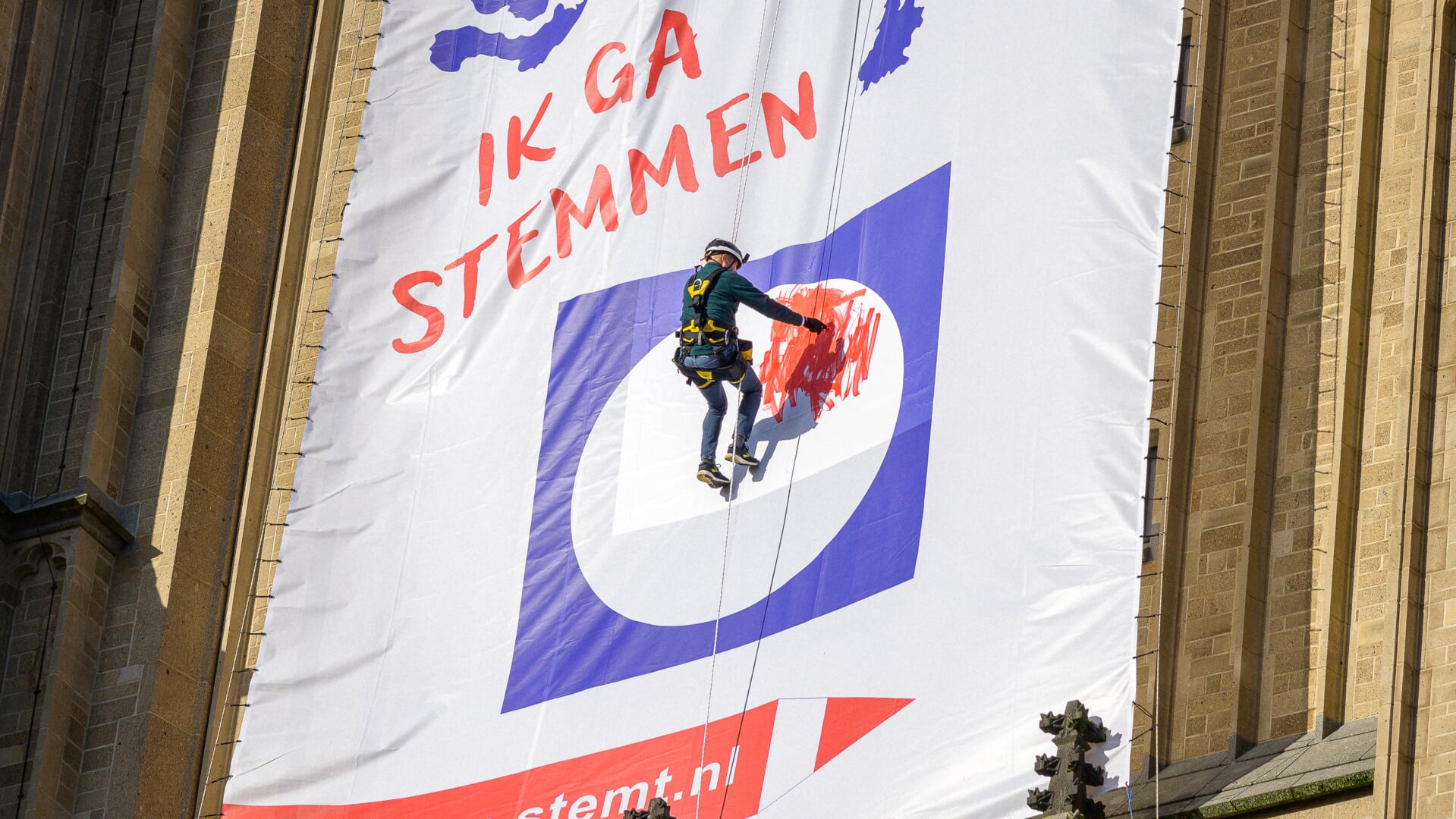 Burgemeester Marcouch hangt in de touwen om zijn stem uit te brengen. Hij kleurt een vakje rood op een groot spandoek dat hangt aan de Eusebiuskerk.