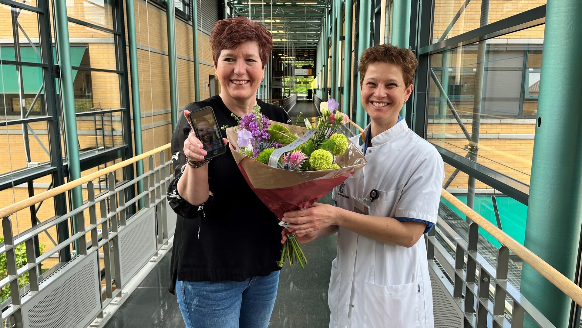 Mevrouw Smeets krijgt bloemen van verpleegkundig specialist Janneke van Aalst-Roes. Op haar telefoon een zorgverlener via de Virtual Clinic-app.