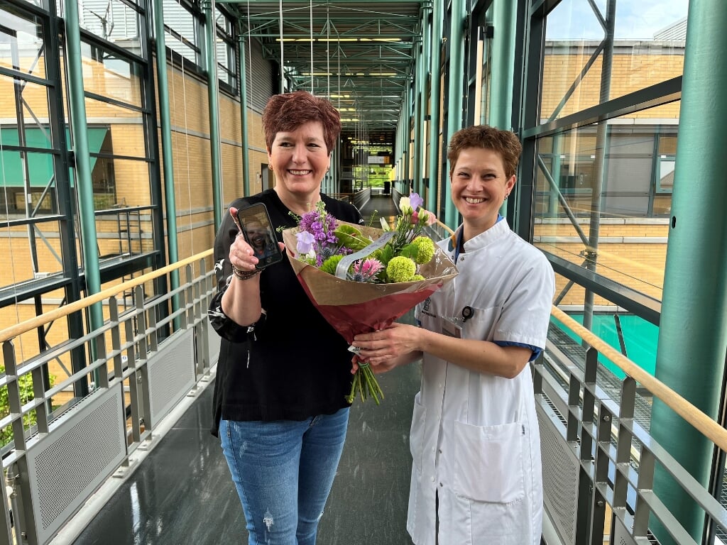 Mevrouw Smeets krijgt bloemen van verpleegkundig specialist Janneke van Aalst-Roes. Op haar telefoon een zorgverlener via de Virtual Clinic-app. 